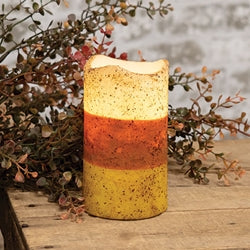 A timer pillar candle with a color block yellow, orange, and beige design, resembling candy corn, with a grungy texture. It is placed on a surface with some foliage at the base.